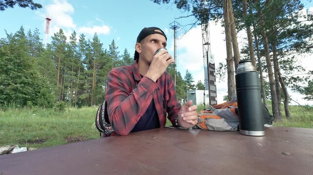 Adult man in backward cap and red plaid shirt sits at weathered picnic table in pine forest clearing, pausing to sip from metal cup beside backpack while looking across sunlit woodland