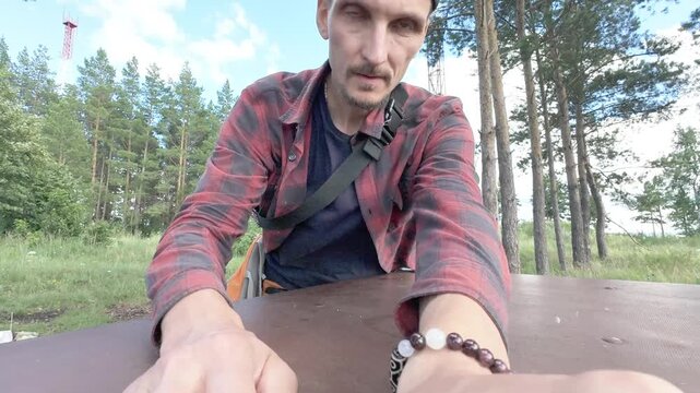 Low angle motion scene of adult man in backward cap and red plaid shirt packing backpack on round picnic table in pine forest clearing with grass, wood texture, and soft natural light