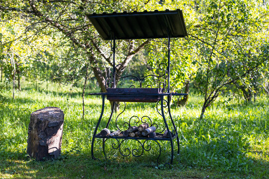 Ornamental metal garden grill stand with stacked firewood and stump in green orchard for outdoor picnic background