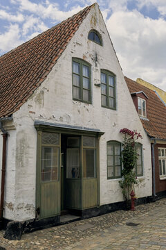 View of a historic white timber-framed house with a red tiled roof, green window frames, and a climbing rose bush on a cobblestone street in Ribe, Denmark.