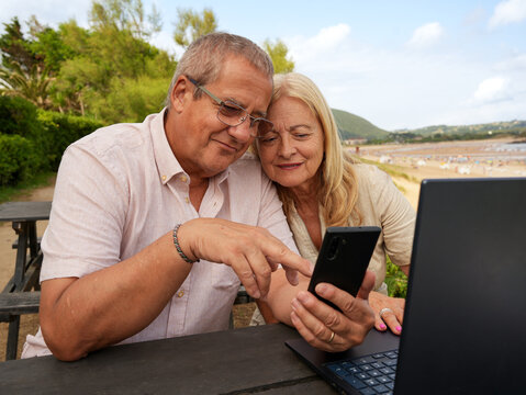 Senior couple use smartphone and laptop at beach cafe