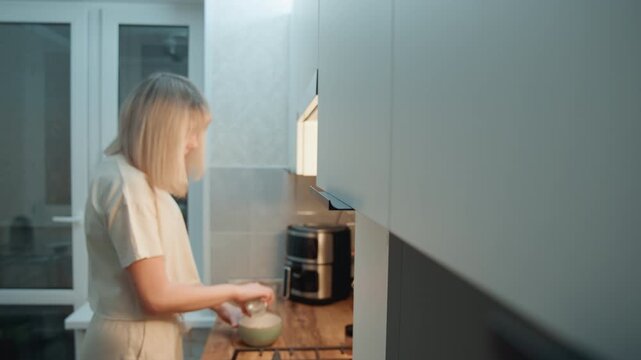 Blonde caucasian woman preparing breakfast in bright kitchen with daylight, mixing ingredients in bowl beside coffee maker, efficient focused pace, modern appliances, tidy counter, purposeful domestic