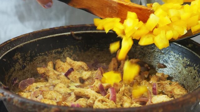 In a deep cast-iron skillet, onions and chicken are simmered in a sauce and spices, with yellow pepper added. Medium shot.