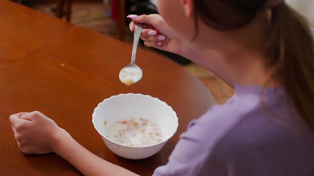 Young woman holds spoon over white bowl of cereal with milk, seated at wooden table wearing purple shirt, morning breakfast scene.