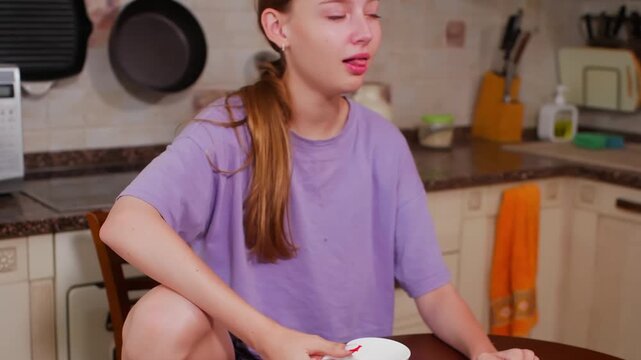Young woman in purple shirt holds and drink from white mug while sitting at table in home kitchen, appearing calm during casual moment.
