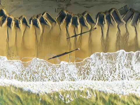 Aerial view of Ellis Beach with unique geometric patterns in the sand, crashing waves, and two people casting long shadows on the shoreline in Cairns, Queensland, Australia.