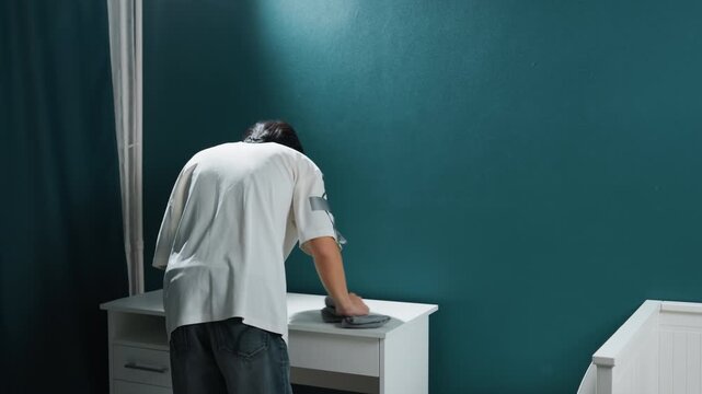 Man cleaning white dresser teal wall methodical homeowner wiping top surface of bedside dresser calm domestic bedroom scene with dark curtain minimal modern furniture overhead light focused hands