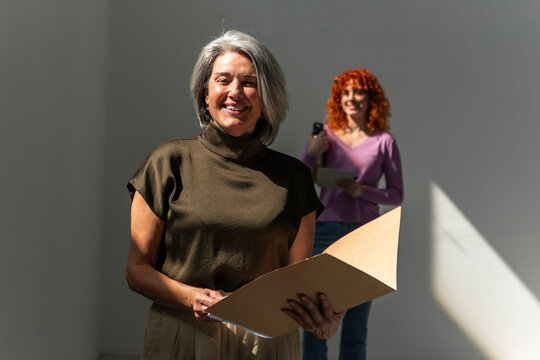 Smiling mature woman holding folder with colleague behind