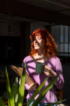 Young businesswoman holding folder and coffee near plant