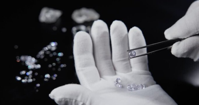 Slow motion jeweler inspecting and sorting loose diamonds on gloved hand in dark studio tabletop.
