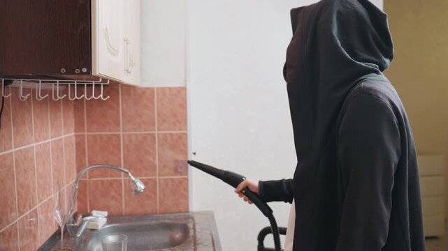Moody kitchen deep cleaning at sink by white homeowner in hoodie using steam nozzle to remove grime from tiles and faucet, domestic chore vibe with soft natural light and focused maintenance action
