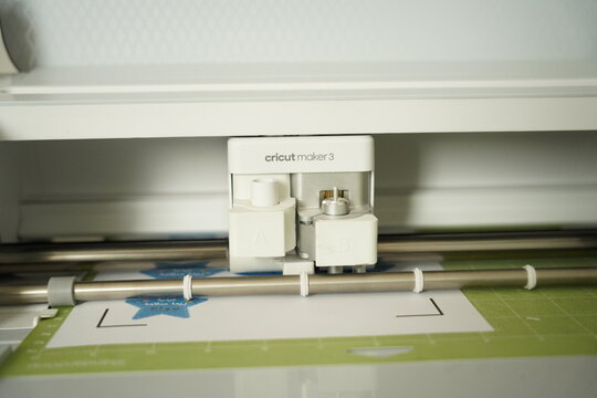 Close-up of a Cricut Maker 3 electronic cutting machine working on a printed craft sheet and cutting mat, showing the tool carriage and blade in a home crafting workspace.