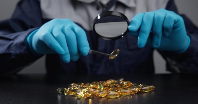 Slow motion close up technician with gloved hands inspecting golden capsules on laboratory table with magnifying glass.