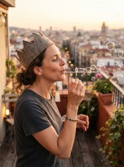Fototapeta premium A middle aged caucasian woman wears a felt crown and blows soap bubbles on a sunlit urban balcony terrace.