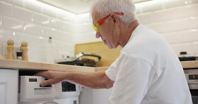 Senior man repairing washing machine in modern kitchen.