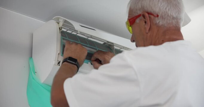 Slow motion senior man inspecting and cleaning air conditioner filter inside home near ceiling and wall.