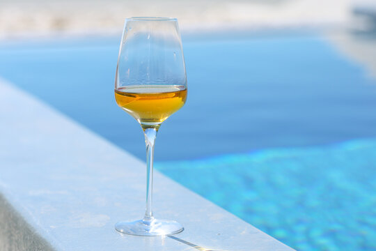 A glass of golden white wine stands on a smooth ledge beside a shimmering blue pool, capturing the essence of summer relaxation and Mediterranean leisure in bright sunlight.