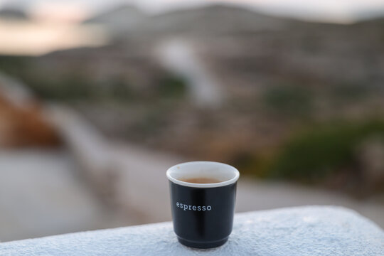 A black espresso cup rests on a white ledge overlooking a blurred rocky landscape, capturing a peaceful moment of coffee enjoyment in natural light.