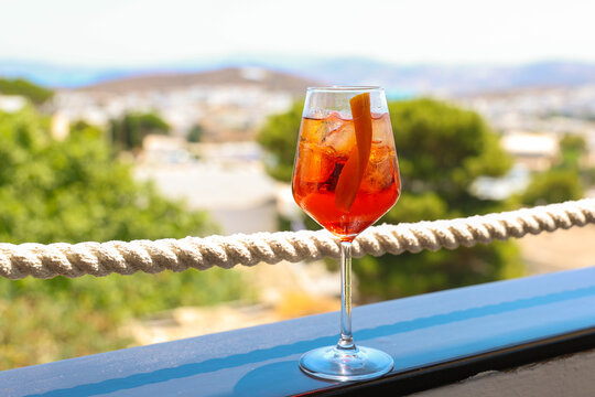 A glass of Aperol spritz with ice and orange peel sits on a terrace railing overlooking trees and hills, symbolizing summer relaxation and Mediterranean charm.