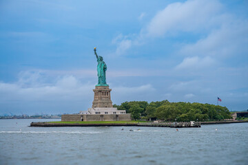 Statue of Liberty on Liberty Island viewed from Hudson River. Copper statue on stone pedestal surrounded by green trees under cloudy sky with rippling water in foreground. © Vadim