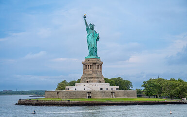 Statue of Liberty on stone pedestal in New York bay. Full height copper figure with torch standing on granite base near calm water under soft light. © Vadim
