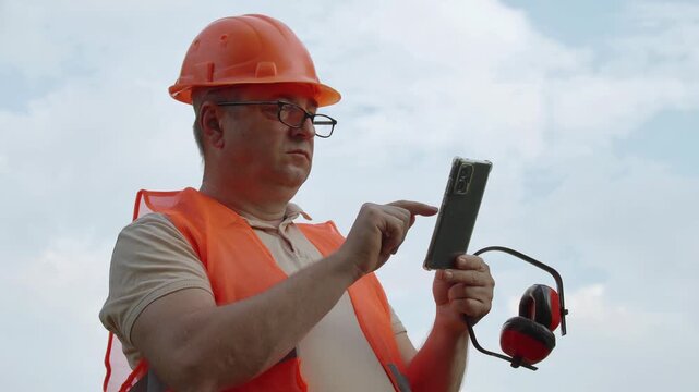 Builder wearing an orange safety vest and hard hat engages with a smartphone while holding earmuffs, positioned outdoors with a cloudy sky in the background, emphasizing safety in construction