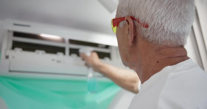 Elderly man spraying and cleaning wall mounted air conditioner inside home with protective drape and close handheld movement.