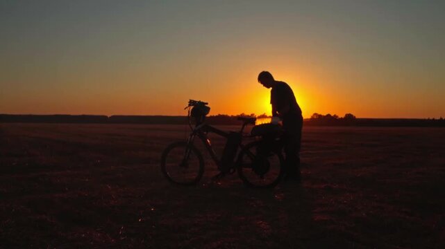Silhouette of a male traveler with an electric bicycle taking off his backpack in a rural field at sunset. Eco-friendly travel and active lifestyle concept.