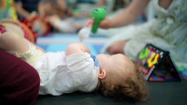 Young baby exploring a green toy frog on a play mat with colorful toys nearby in a nursery setting