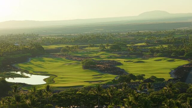 Aerial drone view of luxury golf course glowing at sunrise on Big Island Hawaii with green fairways, tranquil pond, palm groves, black lava fields and volcanic mountain on hazy horizon.