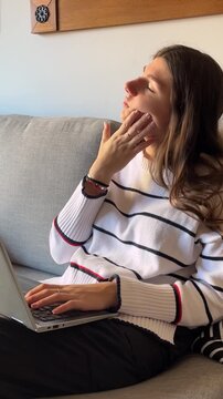Young woman typing on a laptop showing various emotions from concentration to stress and boredom while working or studying from home on a comfortable gray sofa
