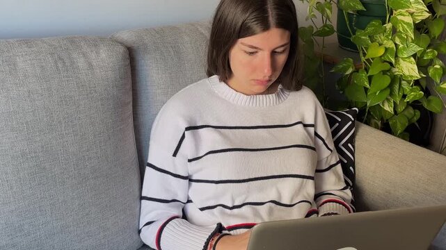 Stressed teenage student working hard on her laptop computer while sitting on a couch at home, showing signs of burnout, fatigue, and frustration from a long day