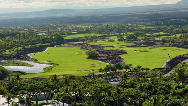 Aerial drone view of a lush green golf course on Big Island Hawaii with sand bunkers, palm trees, a pond and black lava fields stretching toward distant volcanic mountains under soft clouds.