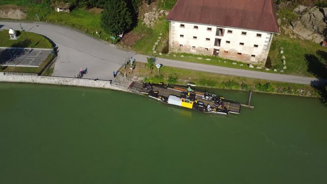 Aerial orbit showing motorcyclists riding onto a small cable ferry on the river Danube near Haibach ob der Donau, Upper Austria, with cyclists waiting at the dock and a historic building on the bank o