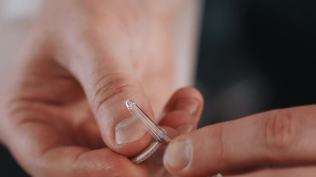 Medical worker readying urgent medication. Detail of hands handling glass ampoule in clinical setting. Intimate view of healthcare worker administering precise medication under subdued lighting