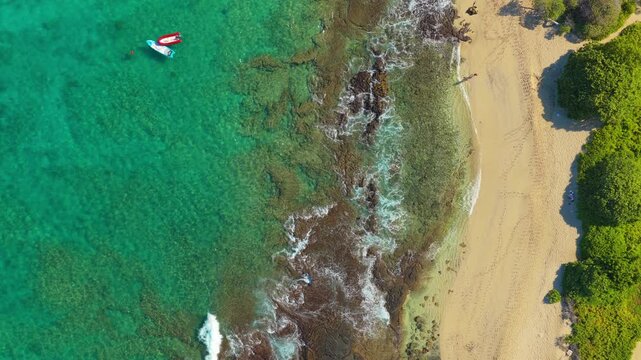 Drone aerial over Big Island Hawaii showing turquoise ocean water waves breaking on volcanic rocky reef moored boats paddleboarders and golden sandy beach lined with green tropical shrubs.