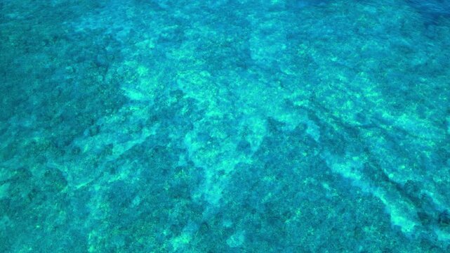 Aerial drone view of crystal clear turquoise ocean water over shallow coral reef along Big Island Hawaii coast. Sunlight ripples across rocky seabed revealing green and blue tropical patterns.