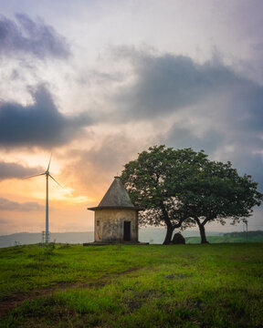 View of a small weathered stone shrine standing beside a large wind turbine and a lush tree in a grassy field under a dramatic sunset sky in India.
