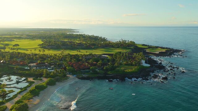Drone aerial view of luxury resort villas and green golf course on lava rock coastline of Big Island Hawaii. Turquoise ocean waves roll onto sandy beach lined with palm trees at golden sunrise.