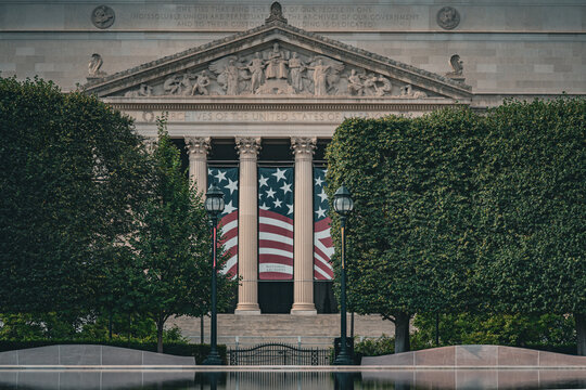 View of the National Archives Building with its neoclassical columns and American flags displayed behind manicured trees and a reflecting pool in Washington, District of Columbia, United States.