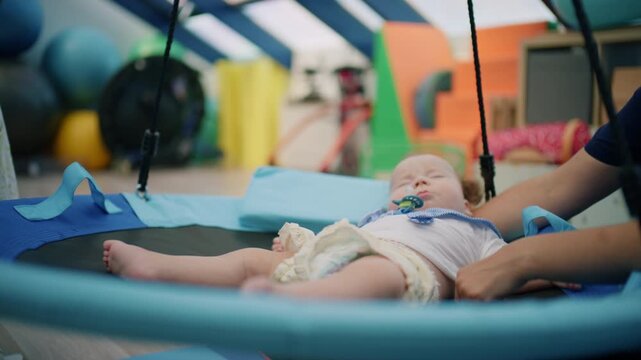 Baby lying on a blue sensory swing during a rehabilitation session, with an adult supervising the therapy