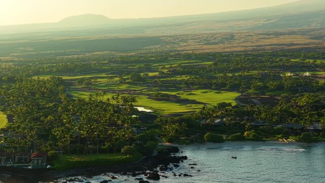 Aerial view of lush green golf course fairways palm groves and luxury homes along the volcanic coastline of the Big Island Hawaii with calm turquoise ocean water glowing in golden sunset light.