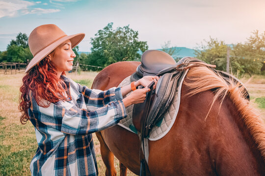 Close-up portrait of a chestnut horse being saddled by a young woman wearing a felt hat at a rural farm. Equestrian lifestyle concept.