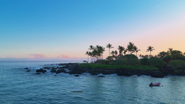 Aerial drone view of tropical Big Island Hawaii coastline at sunset with tall palm trees black lava rock shoreline calm turquoise ocean water pink clouds and small boat moored near green resort.