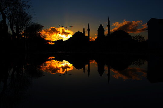 View of Mevlana Museum silhouetted against a vibrant sunset with its reflection visible in a puddle in Karatay, Konya, Turkiye.
