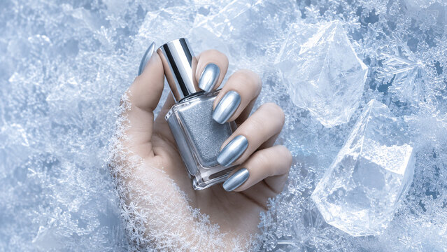 Female hand with metallic silver manicure holding nail polish among ice cubes and frost. Cool winter beauty image for cosmetics, advertising, branding, social media and product presentation.