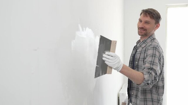 Male construction worker wearing protective gloves and plaid shirt, spreading plaster smoothly across wall surface using professional drywall taping knife during renovation project. Portrait view