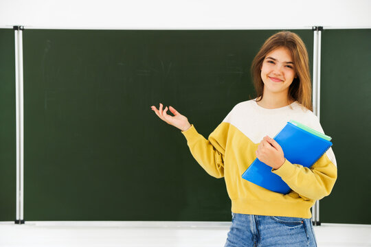 Teenage girl with notebooks pointing at chalkboard in classroom. Space for text