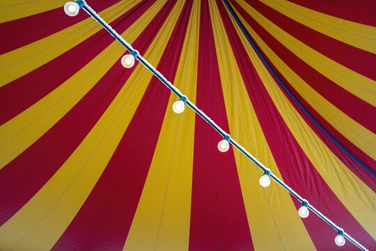 Bright red, yellow striped circus tent ceiling at night. Glowing light bulbs hang on diagonal string covered in frost. Festive carnival marquee canvas glows with warm illumination against cold winter