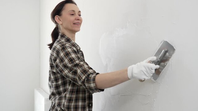 Woman wearing protective gloves and checkered shirt is spreading finishing putty across white wall surface, smoothing damaged areas before painting interior renovation work